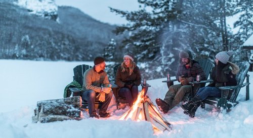 Four people sitting around a campfire in the snow, enjoying drinks and a winter landscape in the background.