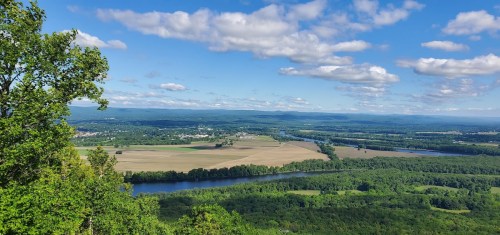 A panoramic view of a lush green landscape with a river, fields, and rolling hills under a blue sky with fluffy clouds.