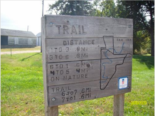 Trail sign displaying distances to various points, including Oak Oak, with a map outline and nearby buildings in the background.