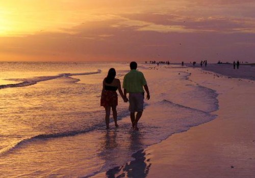 A couple walks hand in hand along a beach at sunset, with gentle waves lapping at their feet.