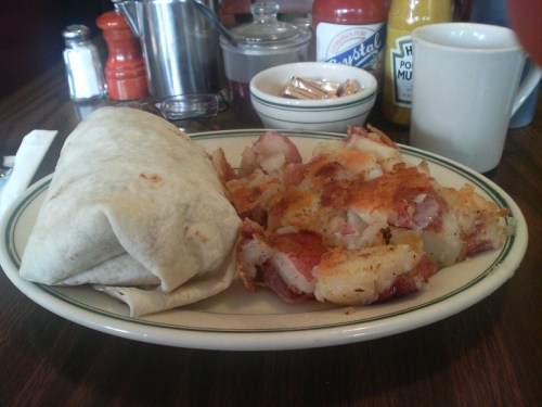 A plate with a burrito and crispy hash browns, accompanied by condiments and a cup on a wooden table.