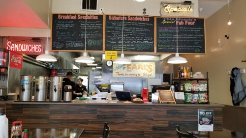 A sandwich shop interior with a menu board, counter, and staff preparing food. Signs highlight specials and offers.