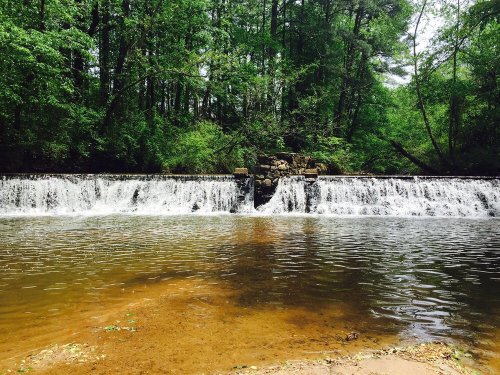 A serene waterfall cascades over a stone barrier into a calm river, surrounded by lush green trees.