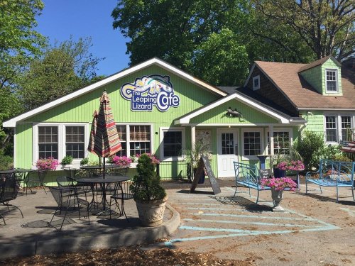A colorful café with outdoor seating, surrounded by greenery and flowers, featuring a sign that reads "The Leaping Lizard Café."