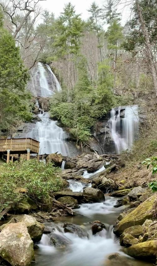 A serene waterfall cascades down rocky cliffs, surrounded by lush greenery and a wooden viewing platform.
