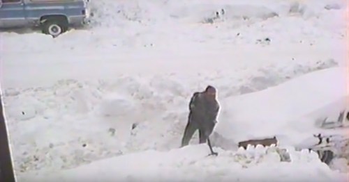 A person shoveling snow in a snowy landscape, with a vehicle partially visible in the background.
