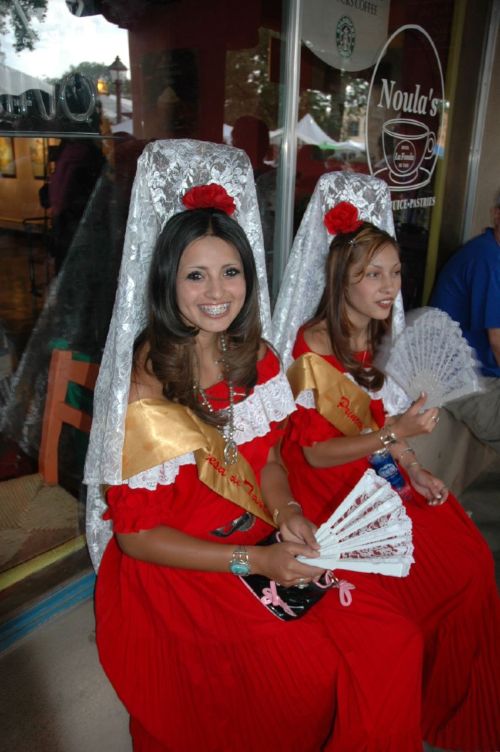 Two women in traditional red dresses with white lace veils, holding fans, seated outside a café.