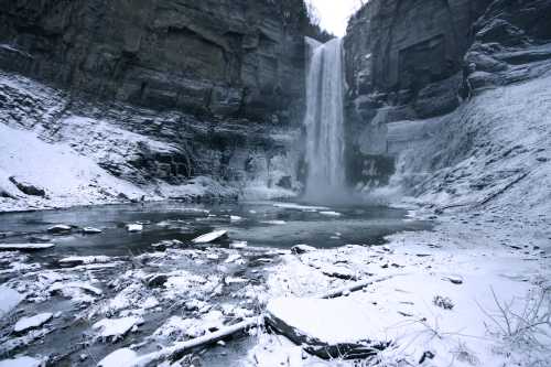 A snowy landscape featuring a tall waterfall cascading into a frozen pool, surrounded by rocky cliffs.