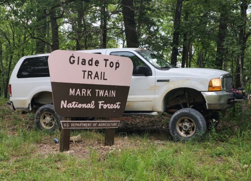 A white SUV parked near a sign for Glade Top Trail in Mark Twain National Forest, surrounded by trees and grass.