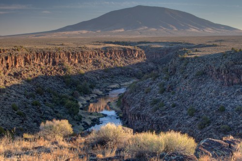 A scenic view of a canyon with a river, framed by rocky cliffs and a distant mountain under a clear sky.
