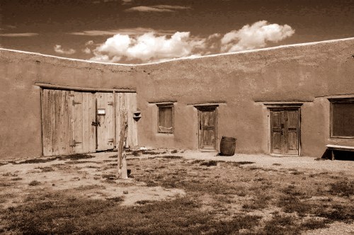 A sepia-toned image of a rustic adobe building with wooden doors and a grassy courtyard under a cloudy sky.