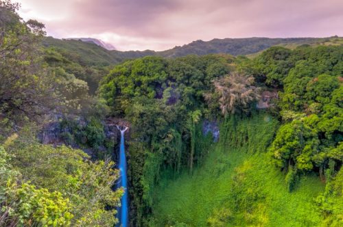A lush green landscape featuring a waterfall cascading into a verdant valley, surrounded by mountains under a cloudy sky.