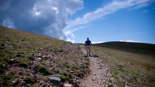 A hiker walks along a rocky trail under a blue sky with clouds, surrounded by grassy hills.