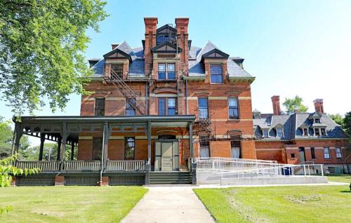 A large, historic brick building with multiple gables, a porch, and fire escapes, surrounded by green grass and trees.