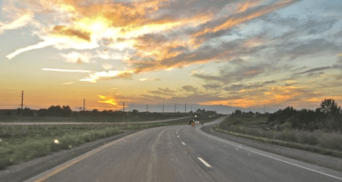 A winding road under a colorful sunset with clouds, surrounded by fields and distant power lines.