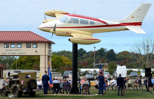 A small plane mounted on a pole at an airport, with a crowd seated nearby, honoring veterans and military service.