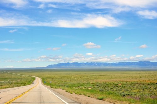 A long, winding road stretches through a vast green landscape under a blue sky with scattered clouds. Mountains are in the distance.