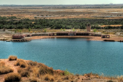 A serene landscape featuring a calm blue lake surrounded by dry land and a rustic building along the shore.