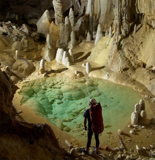 A person stands in a cave, gazing at a turquoise pool surrounded by stalagmites and colorful rock formations.
