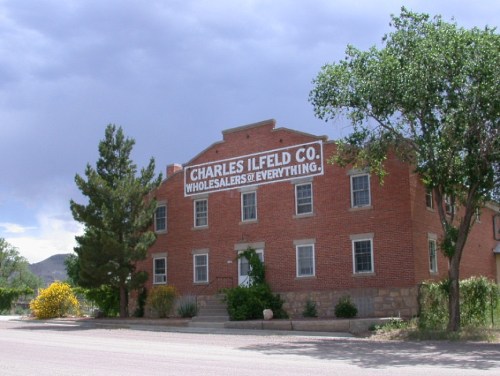 Historic brick building with a sign reading "Charles Ilfeld Co. Wholesalers of Everything," surrounded by trees and shrubs.