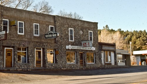 A vintage stone building with storefronts, including a Western wear shop and a cafe, set in a rural landscape.