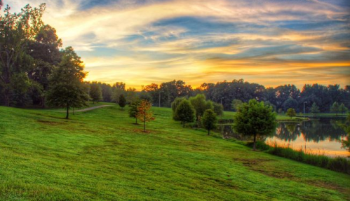 A serene landscape at sunset, featuring a grassy area, trees, and a calm lake reflecting the colorful sky.