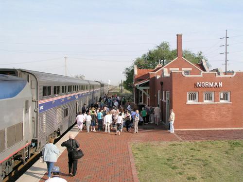 A crowd gathers at the Norman train station as a train arrives, with a brick building labeled "NORMAN" in the background.