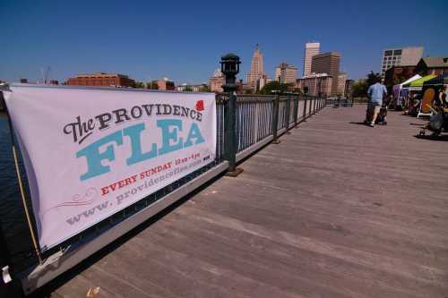 A banner for "The Providence Flea" on a sunny day by the water, with a city skyline in the background.