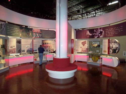 A museum exhibit featuring displays and artifacts, with red lighting and a central pillar. A visitor is exploring the area.