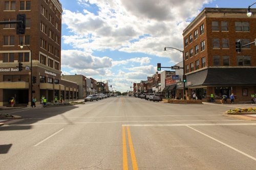 A wide street in a small town, lined with buildings and shops, under a partly cloudy sky. Traffic lights visible.