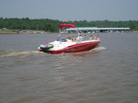 A red and white boat with passengers speeds across a calm lake on a sunny day.