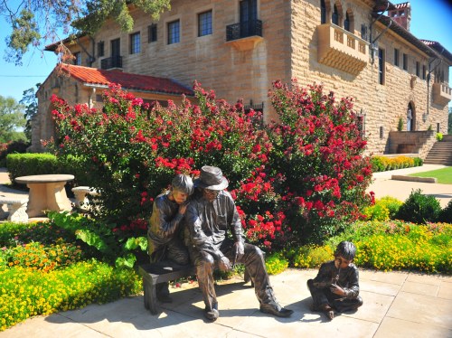 A bronze sculpture of a seated couple with a child nearby, surrounded by vibrant flowers and a historic building.