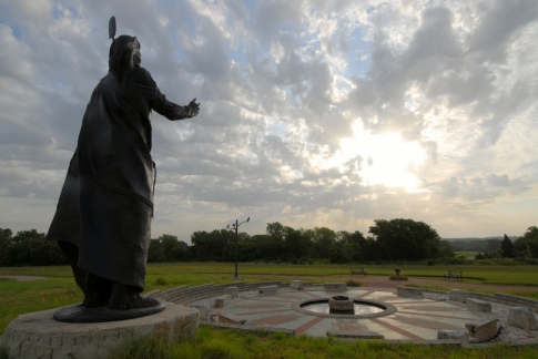 A large statue stands on a grassy hill, overlooking a circular stone structure under a cloudy sky at sunset.