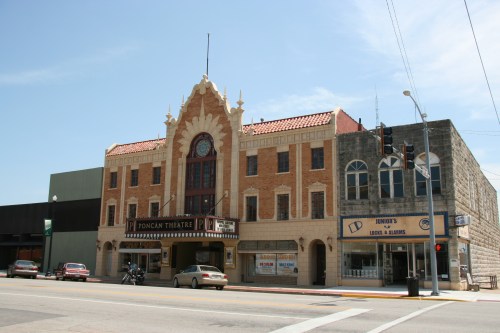 Historic theater building with ornate architecture, featuring a marquee and adjacent shops on a sunny day.