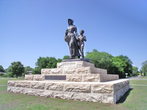 A bronze statue of a woman and a boy stands on a stone pedestal, surrounded by green trees and a clear blue sky.