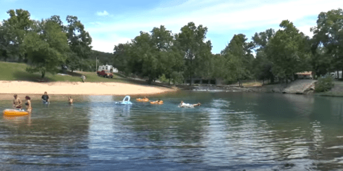 A serene lake scene with children playing in the water, surrounded by trees and a sandy beach in the background.