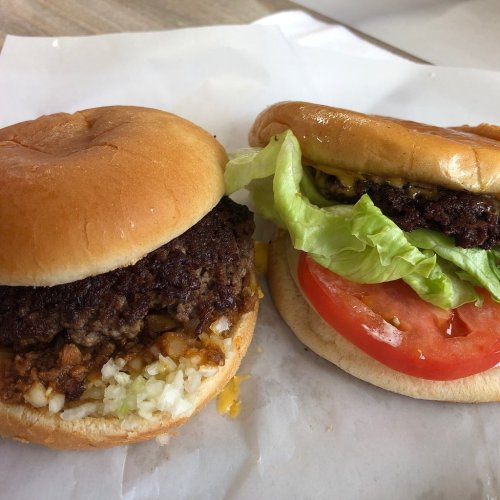 Two hamburger sandwiches on a white surface: one with a beef patty, onions, and cheese; the other with lettuce and tomato.