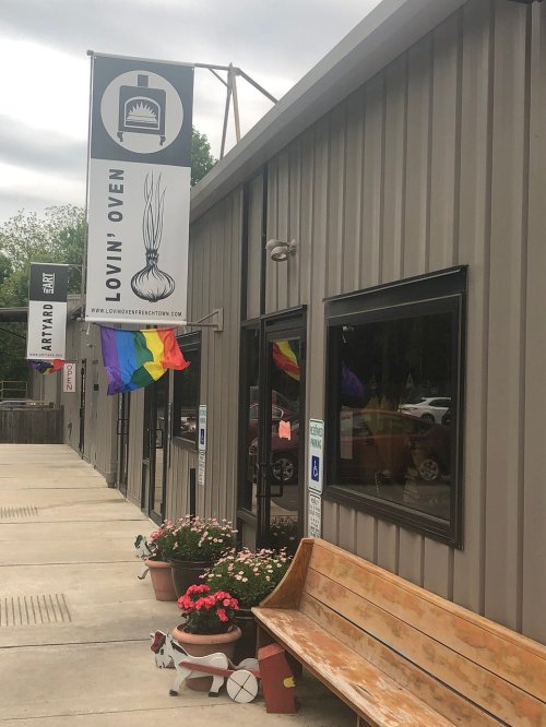 Exterior of a building with a sign reading "Loving Oven," featuring rainbow flags and flower pots by a wooden bench.