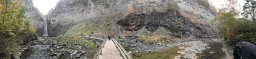 A panoramic view of a waterfall surrounded by steep cliffs and lush greenery, with a wooden bridge in the foreground.