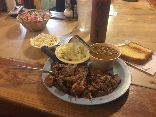 A plate of sliced beef with sides of coleslaw, potato salad, baked beans, and a slice of bread on a wooden table.