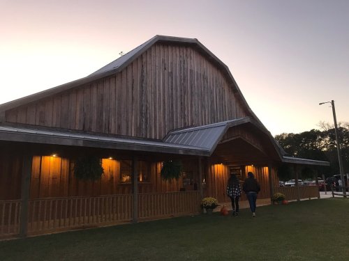 A wooden barn-style building at sunset, with warm lights and people walking towards it.