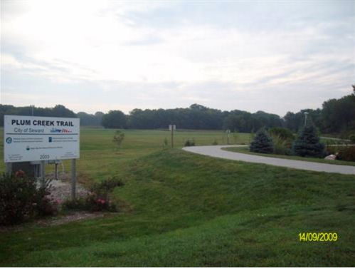 Sign for Plum Creek Trail in a grassy area with a paved path, trees in the background, under a cloudy sky.