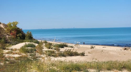 A serene beach scene with gentle waves, sandy dunes, and a clear blue sky, featuring a person walking along the shore.