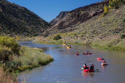 A group of people kayaking on a calm river surrounded by green hills and rocky terrain under a clear blue sky.