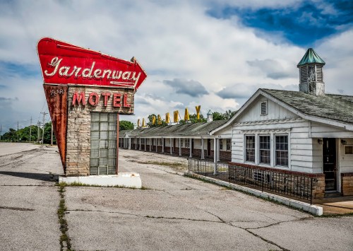 A vintage motel sign stands in front of an empty, weathered motel building under a cloudy sky.