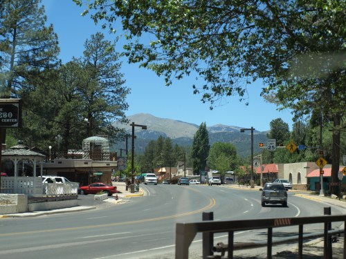 A scenic view of a small town street lined with trees, shops, and mountains in the background on a sunny day.