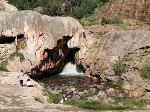A serene landscape featuring a small waterfall flowing from a rocky cave, with people sitting nearby on the bank.