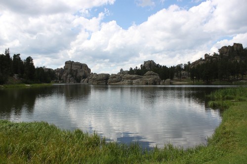 A serene lake surrounded by rocky formations and lush greenery under a cloudy sky. Reflections shimmer on the water.