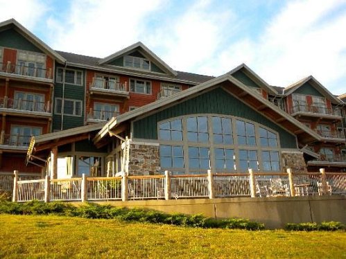 A large, modern lodge with a stone facade and multiple balconies, set against a blue sky. Green grass in the foreground.