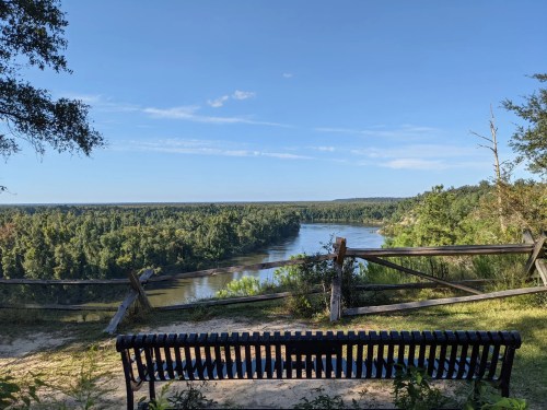 A scenic view of a river surrounded by trees, with a bench in the foreground and a clear blue sky above.